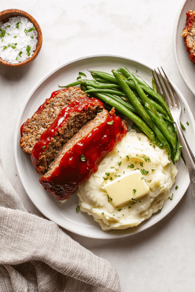 Classic meatloaf slices with mashed potatoes, butter, and green beans on a white plate, comforting and homemade meal