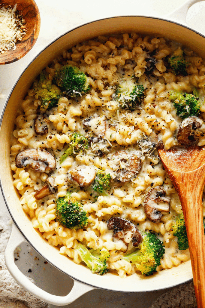 Cheesy one-pot broccoli mac and cheese with mushrooms in a creamy sauce, cooked perfectly in a white pot with a wooden spoon