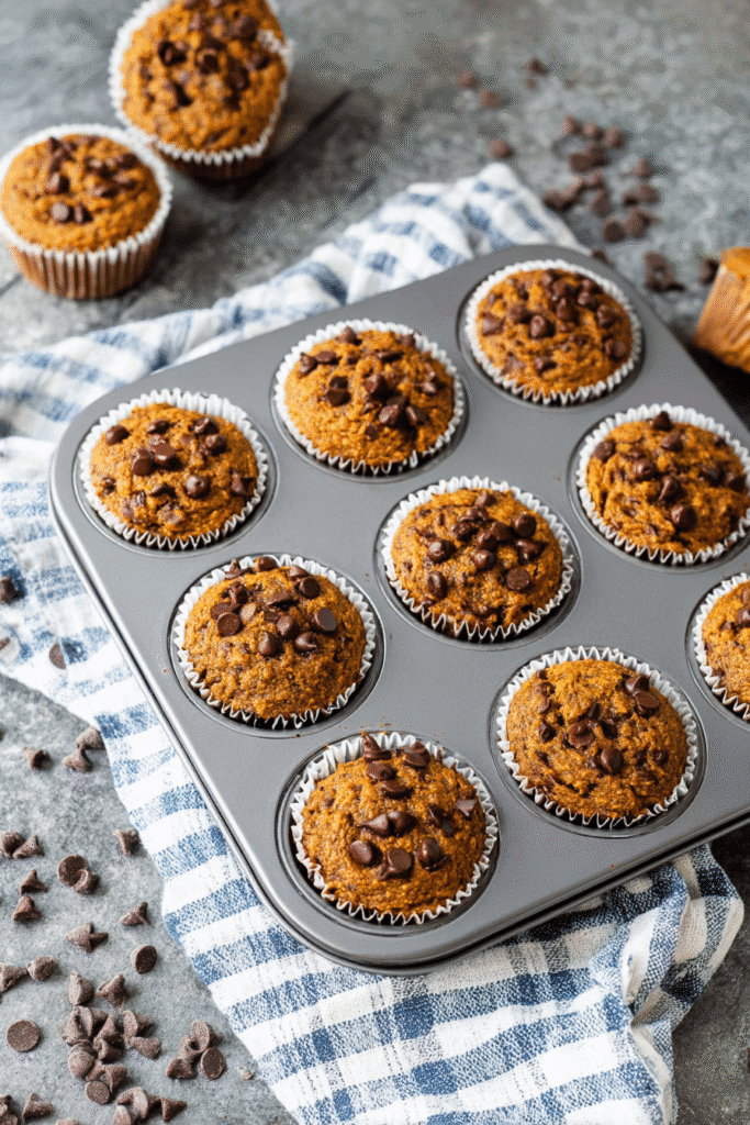 Gluten-free keto pumpkin muffins topped with chocolate chips cooling in a muffin tray on a blue checkered kitchen towel