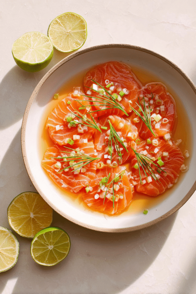 Soy citrus salmon sashimi garnished with green onions and herbs in a light sauce with lime halves on the side