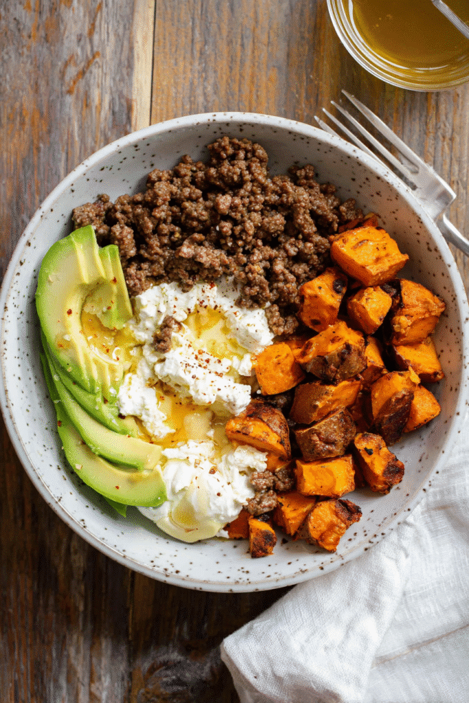 Ground beef bowl with roasted sweet potatoes, avocado, sunny-side egg, and creamy cheese drizzled with hot honey.